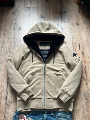 Beige hoodiejacka från Moose Knuckles - Snygg beige jacka från Moose Knuckles med dragkedja framtill och svart foder i huvan. Har två fickor fram och en ikonisk logga på ärmen. Tillverkad i ett tjockare material som känns lyxigt och mjukt. Perfekt för dig som vill ha en stilren och exklusiv look.