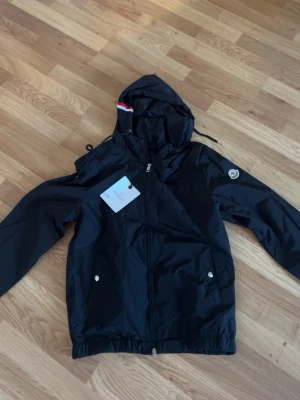 Svart vindjacka från Monclerr - Snygg svart vindjacka från Moncler med huva och dragkedja framtill. Jackan har två fickor med tryckknappar, elastisk nederkant och en Moncler-logga på ärmen. Materialet är lätt och vattenavvisande, perfekt för blåsiga dagar.