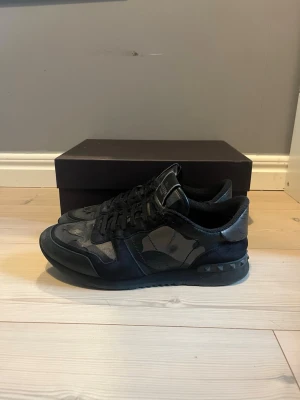 Svarta sneakers från Valentino - Säljer ett par svarta sneakers från Valentino med coola kamouflagemönster och nitar på hälen. Skorna har en låg profil, snörning och är tillverkade i en mix av mocka och textil. Perfekta för dig som gillar exklusiva och stilrena sneakers. Suveräna skor nu inför våren!