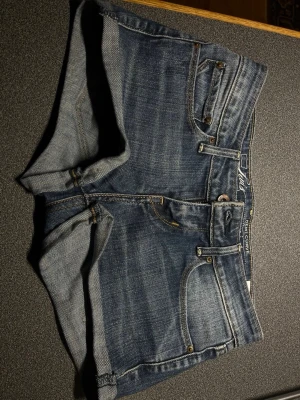Lågmidjade Blå jeansshorts från Levi's - Säljer ett par klassiska blå jeansshorts från Levi's med fem fickor och uppvikta benslut. Shortsen har normal passform och är tillverkade i slitstark bomull. Perfekta för sommaren och har den ikoniska Levi's-detaljen på bakfickan. 