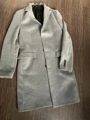 Ljusgrå klassisk ullkappa - Stilren ljusgrå kappa med rak passform och klassisk krage. Kappan har två fickor med lock framtill, knäppning med tre knappar och dekorativa knappar vid ärmslut. Fodrad insida för extra komfort. Perfekt för dig som gillar en tidlös och clean look. Fattas en knapp