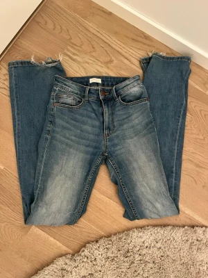 Blå bootcut jeans  - Säljer ett par blå jeans med utsvängda ben. Jeansen har klassisk femficksdesign och snygga slitningar framtill och baktill för en avslappnad look. Perfekta till vardagsstilen och passar till det mesta.