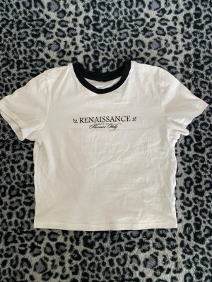 Vit t-shirt med svart text och krage - Säljer en vit t-shirt med svart text 'RENAISSANCE Florence Italy' på bröstet och svart ribbad krage. T-shirten har korta ärmar och en rak passform. Perfekt för dig som gillar enkel men snygg stil. I storlek XS 