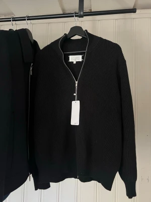 Svart stickad ziptröja Maison Margiela - Svart stickad tröja från Maison Margiela med hel dragkedja framtill och ribbade muddar. Tröjan har lång ärm och den ikoniska vita sömmen i nacken. Perfekt till lager på lager och enkel att styla till olika looks.
