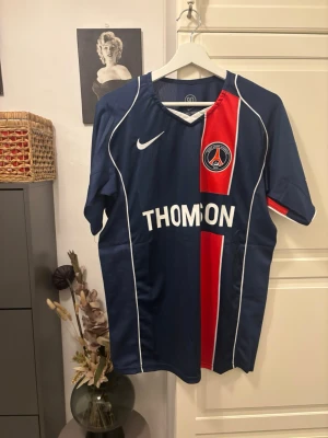 PSG matchtröja Nike blå/röd 2004 home  - Snygg Paris Saint-Germain matchtröja från Nike i mörkblått med röd och vit vertikal rand, THOMSON-tryck på bröstet och PSG-logga. Klassisk sportig design med korta ärmar och v-ringad hals. Perfekt för fotbollsfans!
