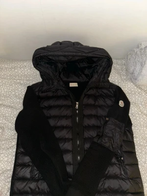 Svart dunväst med huva från Moncler - Snygg svart dunväst från Moncler med huva och dragkedja framtill. Västen har stickade svarta ärmar och klassisk Moncler-logga på ena ärmen. Perfekt för lager-på-lager och streetstyle. Materialet är en kombination av dun och stickat tyg.