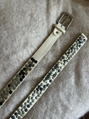 Vitt skärp med nitar - Snyggt vitt skärp i skinn med stora runda nitar i silver och svart längs hela bandet. Från tessie. Skärpet är använt och har därmed lite slitage, vilket syns på bilderna🪩