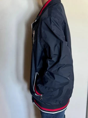 Mörkblå bomberjacka från Santa Barbara - Snygg mörkblå bomberjacka från Santa Barbara Polo & Racquet Club med vita och röda detaljer på krage, ärmslut och nederkant. Jackan har dragkedja, broderad logga och amerikansk flagga på bröstet samt fickor med dragkedja. Tillverkad i lätt, glansigt material.