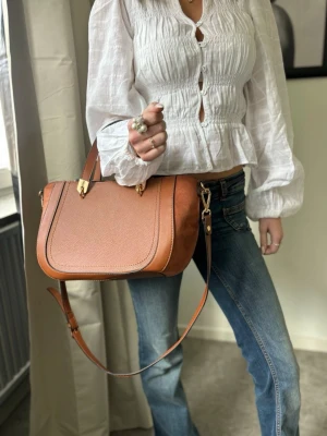 Brun handväska med axelrem - Snygg brun handväska i läderimitation med både handtag och justerbar axelrem. Väskan har guldfärgade metalldetaljer och en stilren, strukturerad form. Perfekt för dig som vill ha en trendig accessoar till din outfit.