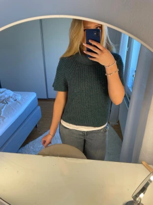 Mörkgrön stickad t-shirttröja - Säljer en mörkgrön stickad t-shirttröja med korta ärmar och hög, ribbad krage. Tröjan har en grovstickad struktur och är perfekt för lager på lager. Märke och material är okänt men den känns mjuk och skön mot huden. Vet ej vilken strl men den sitter bra på mig som har S/ M och är 170. Köptes för 200kr om jag minns rätt , vet inte vilket märke , se defekt på sista bilden