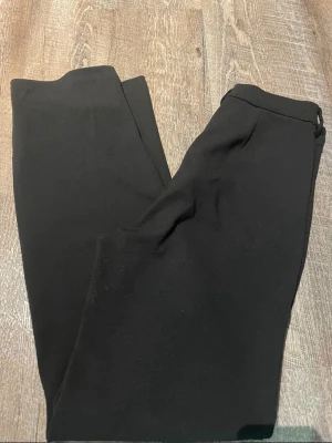 Svarta kostymbyxor från Vero Moda - Snygga svarta kostymbyxor från Vero Moda med klassisk rak passform och diskret dragkedja samt hake i midjan. Perfekta för en clean och stilren look. Materialet känns mjukt och följsamt, vilket gör dem bekväma att bära hela dagen.