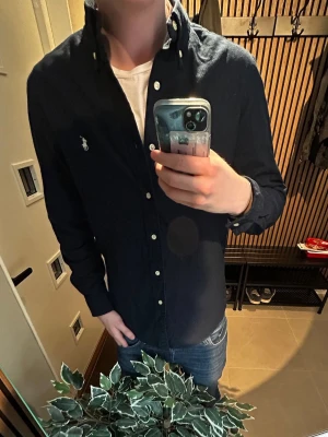 Mörkblå skjorta från Ralph Lauren - Klassisk mörkblå skjorta från Ralph Lauren med button-down krage och vita knappar. Skjortan har lång ärm och den ikoniska broderade loggan på bröstet. Perfekt till jeans för en clean och stilren look.