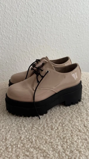 Chunky beige lackloafers med platåsula som nya - Säljer ett par chunky loafers från HM i beige lack med svart grov platåsula. Skorna har snörning med svarta skosnören och metallöljetter. Perfekta för dig som vill ha en edgy och trendig look med extra höjd.