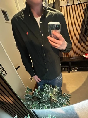 Svart skjorta från Ralph Lauren - Snygg svart skjorta från Ralph Lauren med klassisk krage och knappar framtill. Skjortan har lång ärm och den ikoniska röda loggan broderad på bröstet. Perfekt till jeans för en stilren look. Skriv vid minsta fundering 💭 