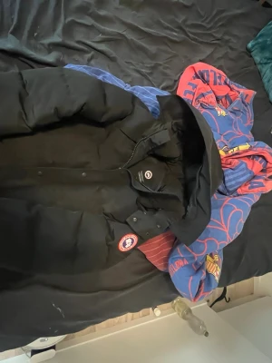 Svart dunjacka från Canada Goose - Säljer en svart dunjacka från Canada Goose med avtagbar huva och klassisk logga på ärmen. Jackan har en fluffig pälskant till huvan och är perfekt för kalla vinterdagar. Stängs med dragkedja och knappar framtill.