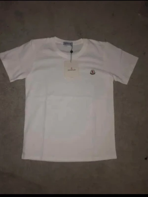 Vit Moncler t-shirt i bomull helt ny - Säljer en clean vit t-shirt från Moncler med klassisk rund hals och korta ärmar. T-shirten har Monclers ikoniska logga broderad på bröstet och är gjord i mjuk bomull för skön känsla. Perfekt till jeans eller shorts för en enkel men snygg look.