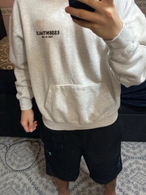 Grå Essentials Fear of God hoodie - Snygg ljusgrå hoodie från Essentials Fear of God med svart logga på bröstet. Klassisk känguruficka framtill och stor huva. Tillverkad i mjukt bomullsmaterial som känns riktigt skönt mot huden. Perfekt för en avslappnad och trendig streetwear-look.