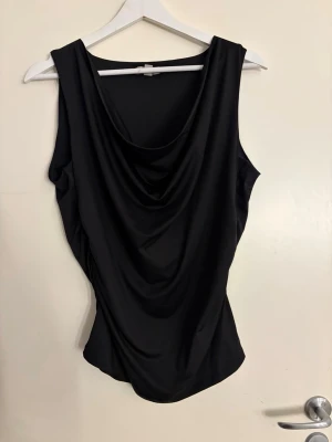 Svart draperad ärmlös topp - Snygg svart ärmlös topp med draperad front och rund halsringning. Toppen har en elegant och stilren look, perfekt att matcha med både jeans och kjol. Materialet känns mjukt och följsamt mot kroppen. Storlek L aldrig använd