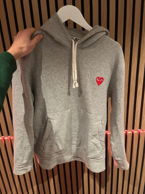 Grå Comme des Garçons Play hoodie - Säljer en grå hoodie från Comme des Garçons Play med det ikoniska röda hjärtat med ögon broderat på bröstet. Hoodien har huva med vita dragsnören och en stor magficka. Tillverkad i mjuk bomullsblandning, perfekt för en chill och stilren look.