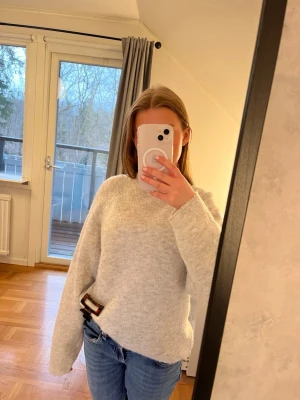 Ljusgrå stickad tröja med hög hals - Mysig ljusgrå stickad tröja med hög hals och avslappnad passform. Tröjan har långa ärmar och ribbstickade muddar vid ärmslut och nederkant. Perfekt för kalla dagar och enkel att matcha med jeans eller kjol.