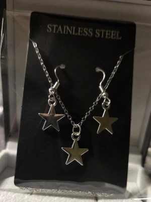Smyckeset med stjärnhängen i stål - Snyggt smyckeset i rostfritt stål med halsband och örhängen, alla med matchande silverfärgade stjärnhängen. Kedjan är tunn och elegant, och stjärnorna ger en cool och trendig look. Perfekt för dig som gillar stilrena accessoarer med lite edge.