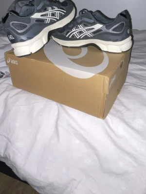 ASICS grå sneakers med vita detaljer - Säljer ett par snygga ASICS sneakers i mörkgrå färg med vita detaljer och logga på sidan. Skorna har en låg profil, mesh och syntetmaterial i ovandelen samt en vit, robust sula. Perfekta för dig som gillar sportig stil och komfort. Skorna är använda ett få tal gånger. Obs att mer bilder kan skickas i chatten.