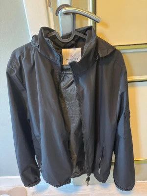 Svart vindjacka från Moncler - Snygg svart vindjacka från Moncler med dragkedja framtill, huva och två fickor med dragkedja. Jackan har en minimalistisk design och elastisk nederkant för skön passform. Perfekt för blåsiga dagar och enkel att matcha med olika outfits.