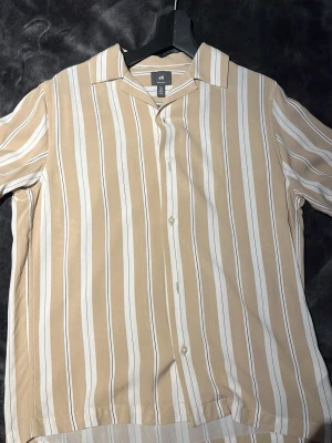 Beige och vit randig skjorta H&M - Säljer en beige och vit randig skjorta från H&M i regular fit. Skjortan har klassisk krage, knäppning framtill och korta ärmar. Perfekt för dig som gillar en avslappnad men ändå stilren look.