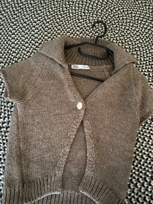 Brun stickad kortärmad kofta Zara - Kortärmad stickad kofta från Zara i en mjuk brun nyans. Koftan har en bred krage, en dekorativ knapp framtill och ribbade muddar vid ärmslut och nederkant. Perfekt att slänga över en topp för en chill look.