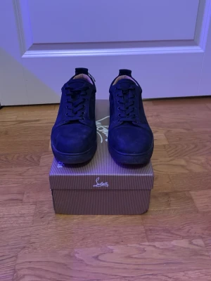 Svarta sneakers från Christian Louboutin - Stilrena svarta sneakers från Christian Louboutin i mocka med klassisk röd sula. Lågt skaft och svarta skosnören. Kommer med originalkartong, dustbag och äkthetskort. Perfekta för dig som vill ha en lyxig och tidlös look.