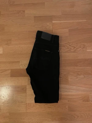 Svarta slim jeans från Nudie Jeans - Säljer ett par svarta slim jeans från Nudie Jeans med klassisk femficksdesign och silverfärgade nitar. Jeansen har en smal passform och är tillverkade i stretchigt denimtyg för extra komfort. Perfekta för en clean och stilren look.