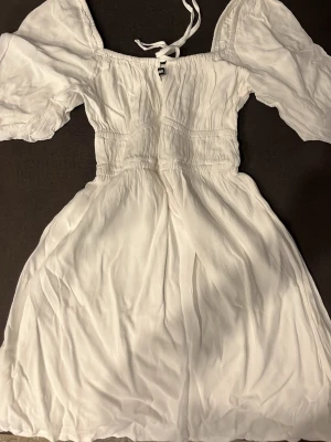 Vit klänning - Säljer en vit klänning från BikBok i storlek XS. Klänningen har puffiga ärmar, smockad överdel och knytband vid halsen. Materialet är lätt och flowy, perfekt för en somrig look. Passar dig som gillar romantiska och trendiga klänningar.