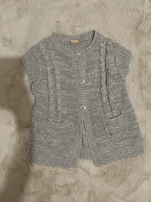 Grå stickad väst från Cherokee - Snygg grå stickad väst från Cherokee med korta ärmar, kabelstickade detaljer framtill och två små fickor. Stängs med tre stora knappar och har ribbade kanter. Perfekt att slänga över en långärmad tröja för extra stil.