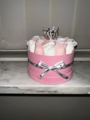 Rosa presentbox med rosor och krona - Säljer en rund rosa presentbox dekorerad med vita och ljusrosa rosor, ett band runt och en glittrig liten krona på toppen. Perfekt som dekoration eller gåva till någon speciell.