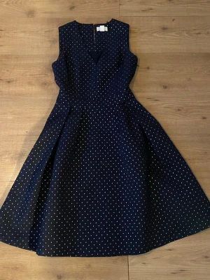Mörkblå prickig ärmlös klänning - Snygg mörkblå klänning från Lindex med vita prickar, v-ringad hals och ärmlös design. Kjolen har en utsvängd form som ger en klassisk siluett. Perfekt för dig som gillar retrovibbar. Klänningen har flera tunna lager av silke i svart 🫶