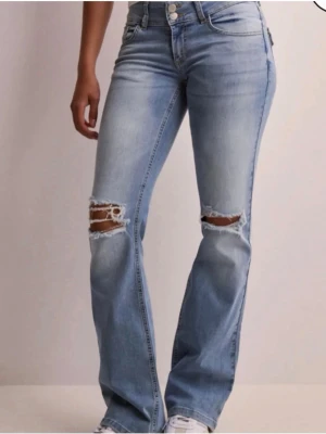 Ljusblå low waist jeans med hål - Säljer ett par ljusblå bootcut jeans med slitna hål över knäna. Jeansen har låg midja, dragkedja och knappstängning med en snygg tvätt.  