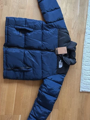 Blå pufferjacka från The North Face - Snygg blå pufferjacka från The North Face med svarta axelpartier och logga på bröstet och ryggen. Jackan har dragkedja framtill, två sidofickor med dragkedja och hög krage. Perfekt för kalla vinterdagar och riktigt trendig stil. Man får även med en gratis vara som en liten present🤯🔥😘 den är i storlek l men sitter mer som s m
