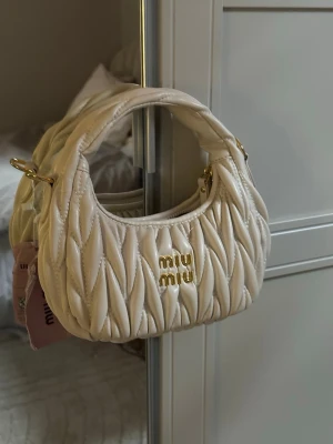 Vit miu miu väska - Supersnygg vit quiltad handväska från Miu Miu med guldfärgade detaljer och logga framtill. Väskan är oanvänd. Väskan har en rundad form, kort handtag och medföljande axelrem. 