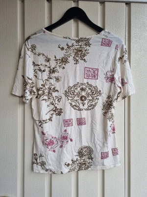 Vit t-shirt med blommigt mönster - Vit t-shirt med korta ärmar och blommigt asiatiskt mönster. Köpt i second hand butik och använts en gång. Fantastiskt fin tröja men säljer pga att det inte riktigt är mina färger 
