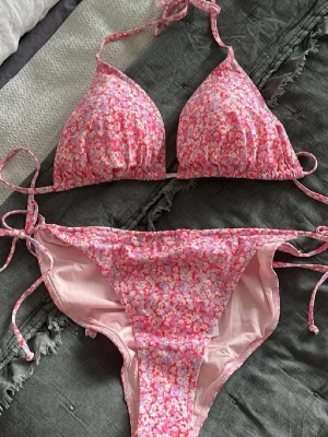 Rosa blommig bikini med knyt - Supersöt bikini i rosa toner med blommigt mönster. Triangeltopp och trosa med knyt i sidorna för justerbar passform. Perfekt för strandhäng och pooldagar. Materialet känns mjukt och följsamt mot huden.