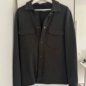 Mörkgrön overshirt från Dressmann - Stilren mörkgrön overshirt från Dressmann med klassisk krage och två stora bröstfickor med lock. Skjortan har långa ärmar och knäpps med tryckknappar framtill. Perfekt att bära som lager över en t-shirt. Hör av er vid funderingar ✅