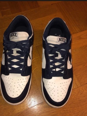 Nike Dunk Low blå/vit sneakers - Snygga Nike Dunk Low sneakers i blått och vitt med klassisk grå Swoosh på sidan. Skorna har perforerad tåbox för ventilation, låga skaft och är tillverkade i läder med textildetaljer. Perfekta för dig som gillar streetwear och sportig stil.