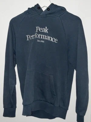 Mörkblå hoodie Peak Performance - Mörkblå hoodie från Peak Performance med vit broderad logga och text 'Est. 1986' på bröstet. Klassisk känguruficka framtill, ribbade muddar och huva med dragsko. Perfekt för en avslappnad och sportig stil.