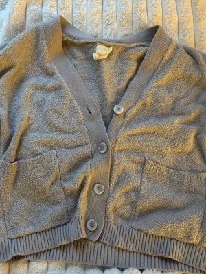 Grå cropped kofta med knappar - Mysig grå cropped kofta med tre knappar framtill och två stora fickor. Koftan har ribbade muddar och är tillverkad i ett mjukt, frottéliknande material. Perfekt för lager på lager och chill dagar.