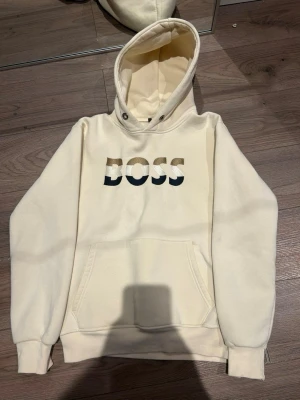 Vit hoodie från BOSS med logga - Snygg vit hoodie från BOSS med stor broderad logga i beige, vitt och svart på bröstet. Hoodien har en klassisk känguruficka framtill, ribbade muddar och justerbar huva. Tillverkad i mjukt bomullsmaterial som känns skönt mot huden.