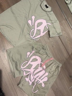 Beige set med rosa tryck - Snyggt tvådelat set i beige med matchande t-shirt och shorts. Båda delarna har ett stort rosa grafiskt tryck framtill och på ryggen. Shortsen har resår i midjan och rosa dragsko. Materialet ser ut att vara mjukt och bekvämt, perfekt för en avslappnad stil.