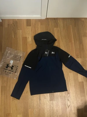 Under Armour marinblå vindjacka - Snygg marinblå vindjacka från Under Armour med svarta detaljer och huva. Jackan har hel dragkedja framtill, reflekterande detaljer på ärmarna och logga på bröstet. Tillverkad i lätt och funktionellt material som passar perfekt för aktiva dagar.