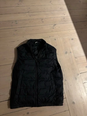 Mörkblå dunväst med dragkedja - Mörkblå dunväst med hög krage och dragkedja framtill. Västen har två sidofickor och quiltad design som ger en sportig look. Perfekt att slänga över en hoodie eller tröja för extra värme. Materialet är glansigt och känns lätt.