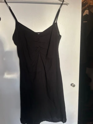 Svart omlottklänning med smala band - Svart klänning med smala axelbandl. Klänningen har en lätt rynkning vid brösten och är i en enkel, rak modell som slutar strax ovanför knät. Perfekt för dig som gillar minimalistisk stil.