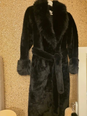 Svart fuskpäls-kappa från Paris - Lyxig svart fuskpäls-kappa från Yu & Me Paris med bred krage, markerade muddar och knytskärp i midjan. Kappan är lång och har en elegant siluett som ger en riktigt trendig look. Perfekt för dig som vill sticka ut med en statement piece i garderoben. Aldrig använd